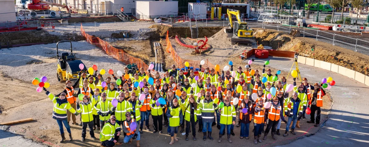Un grand groupe d'ouvriers du bâtiment portant des gilets de sécurité et des casques se tient en cercle sur un chantier, tenant des ballons colorés et posant pour une photo de groupe près de la nouvelle balançoire Sky Swings de Wilderness Explorers à Disneyland Paris.