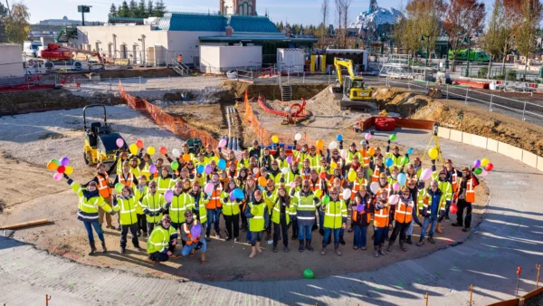 Un grand groupe d'ouvriers du bâtiment portant des gilets de sécurité et des casques se tient en cercle sur un chantier, tenant des ballons colorés et posant pour une photo de groupe près de la nouvelle balançoire Sky Swings de Wilderness Explorers à Disneyland Paris.