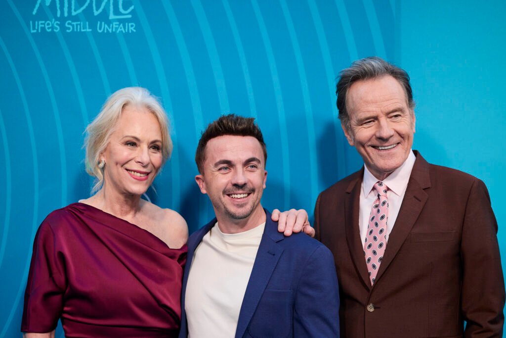 Trois personnes posent ensemble, souriantes, devant une toile de fond bleue lors de la manifestation de l'Avant-première mondiale. Le texte est partiellement visible dans le coin supérieur gauche.