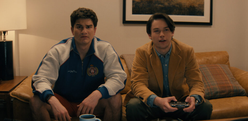 Deux jeunes hommes sont assis sur un canapé ; l'un a l'air confus tandis que l'autre, qui tient une manette de jeu, semble concentré - parfait pour une soirée de streaming Disney+ ou une Nouvelle Comédie inspirée par Mindy Kaling.