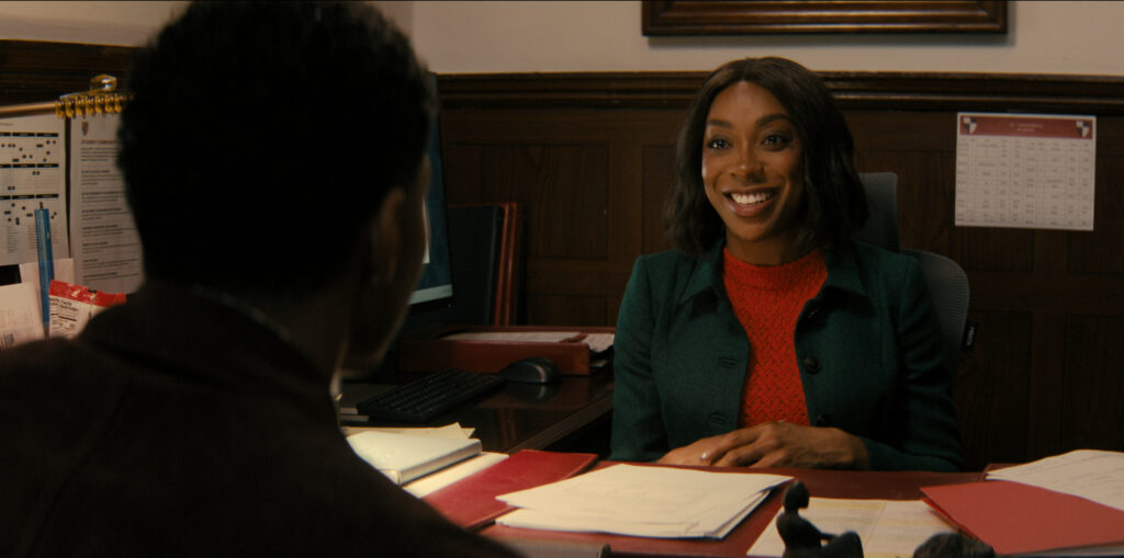 Une femme vêtue d'une veste verte est assise à un bureau avec de la paperasse, souriant à un homme assis en face d'elle dans un environnement de bureau, créant une scène qui rappelle une comédie de Mindy Kaling que l'on pourrait trouver sur Disney+.