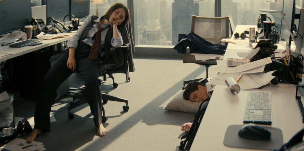 Deux personnes en tenue de travail sont endormies dans un bureau moderne ; l'une est affalée sur une chaise, l'autre est allongée sur le sol avec un oreiller - une scène qui semble tout droit sortie d'un épisode de Not Suitable For Work avec Mindy Kaling.