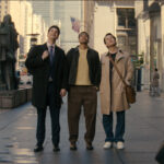 Trois hommes se tiennent sur un trottoir d'une ville animée, regardant vers le haut, tandis que des gens passent et que de grands immeubles s'élèvent à l'arrière-plan, créant une scène digne d'une comédie que vous pourriez trouver sur Disney+.