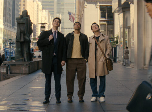 Trois hommes se tiennent sur un trottoir d'une ville animée, regardant vers le haut, tandis que des gens passent et que de grands immeubles s'élèvent à l'arrière-plan, créant une scène digne d'une comédie que vous pourriez trouver sur Disney+.