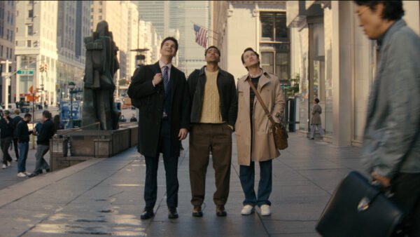 Trois hommes se tiennent sur un trottoir d'une ville animée, regardant vers le haut, tandis que des gens passent et que de grands immeubles s'élèvent à l'arrière-plan, créant une scène digne d'une comédie que vous pourriez trouver sur Disney+.
