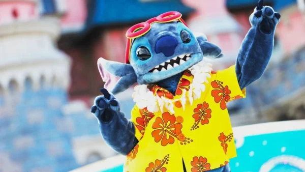 À l'occasion des Stitch Days à Disneyland Paris, une personne déguisée en Stitch de la série Disney "Lilo & Stitch" porte une chemise jaune à fleurs, des lunettes de soleil rouges et un lei, et pose les bras levés devant un château à l'arrière-plan flou.