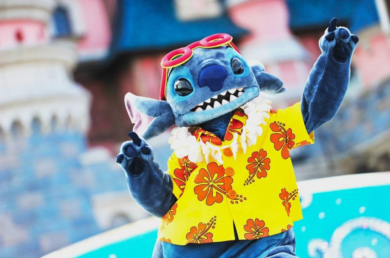 À l'occasion des Stitch Days à Disneyland Paris, une personne déguisée en Stitch de la série Disney "Lilo & Stitch" porte une chemise jaune à fleurs, des lunettes de soleil rouges et un lei, et pose les bras levés devant un château à l'arrière-plan flou.