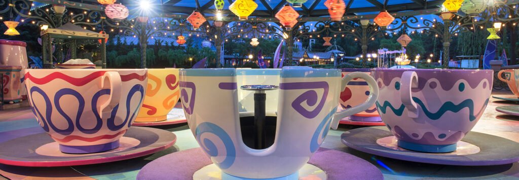 Un manège coloré sur le thème des tasses à thé, avec des tasses et des soucoupes surdimensionnées sous des lanternes suspendues, ravit les visiteurs de Disneyland Paris, dans un parc d'attractions à la tombée de la nuit.