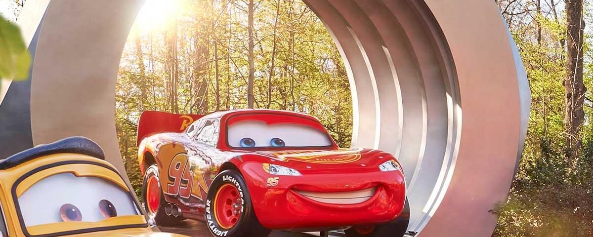 Deux statues de Cars, Luigi et Lightning McQueen, sont exposées sur des rampes devant une grande structure circulaire, alors que la lumière du soleil traverse les arbres. C'est l'endroit idéal pour ceux qui arrivent par la Navette Disneyland ou les Transports Disneyland Paris.