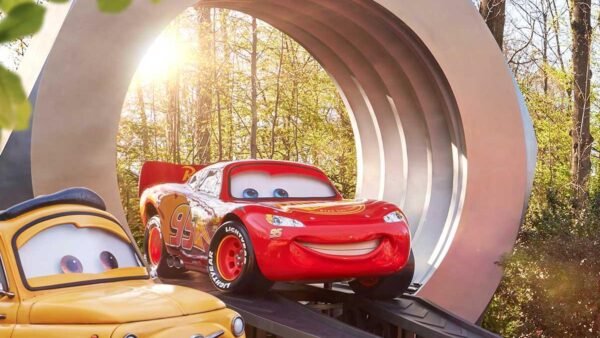 Deux statues de Cars, Luigi et Lightning McQueen, sont exposées sur des rampes devant une grande structure circulaire, alors que la lumière du soleil traverse les arbres. C'est l'endroit idéal pour ceux qui arrivent par la Navette Disneyland ou les Transports Disneyland Paris.