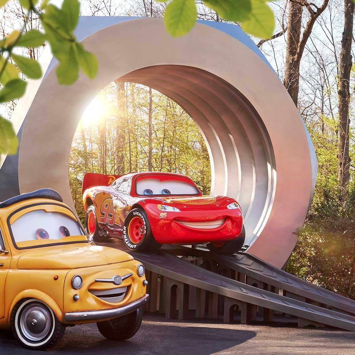 Deux statues de Cars, Luigi et Lightning McQueen, sont exposées sur des rampes devant une grande structure circulaire, alors que la lumière du soleil traverse les arbres. C'est l'endroit idéal pour ceux qui arrivent par la Navette Disneyland ou les Transports Disneyland Paris.