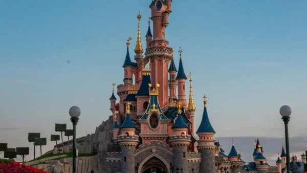 Un grand château de conte de fées rose et bleu, rappelant Disneyland Paris, avec des tours à tourelles, se dresse au centre d'un parc paysager sous un ciel bleu clair.