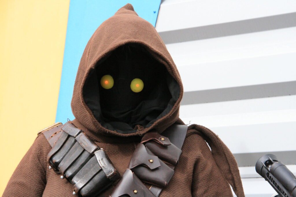 Une personne vêtue d'un costume à capuche marron avec des yeux jaunes brillants, portant un bandolier avec des pochettes, se tient devant un arrière-plan coloré et rayé au magasin pop-up Star Wars de King Jouet.