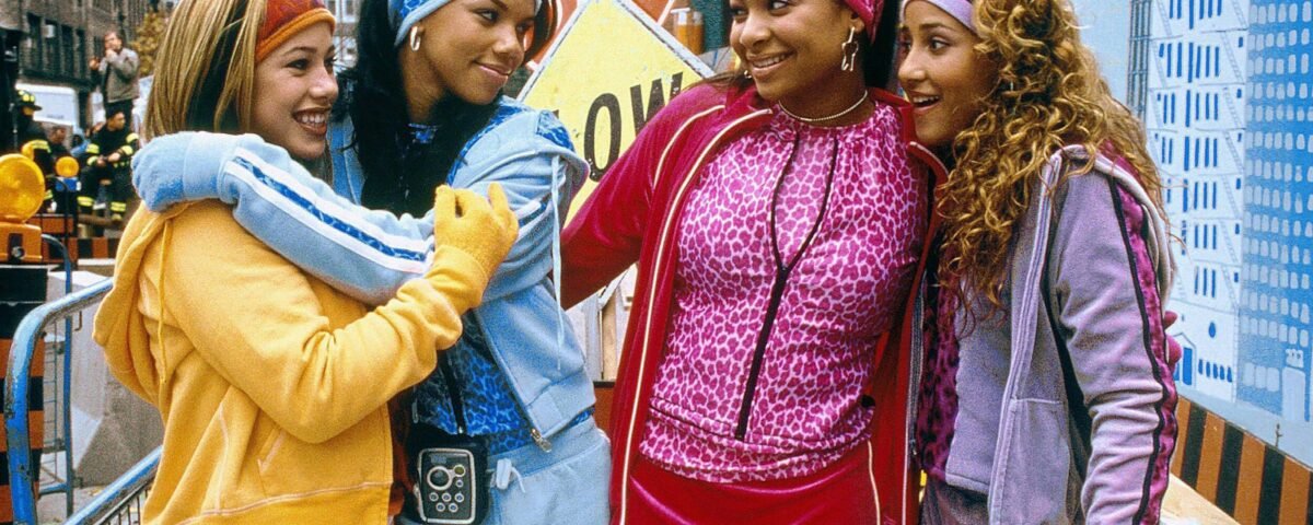 Quatre jeunes femmes vêtues de survêtements colorés se tiennent ensemble, souriantes, dans une rue de la ville, avec des panneaux de construction et des bâtiments en arrière-plan - canalisant l'énergie vibrante des Cheetah Girls d'un film classique de Disney Channel 4e.