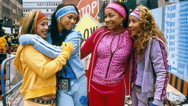 Quatre jeunes femmes vêtues de survêtements colorés se tiennent ensemble, souriantes, dans une rue de la ville, avec des panneaux de construction et des bâtiments en arrière-plan - canalisant l'énergie vibrante des Cheetah Girls d'un film classique de Disney Channel 4e.