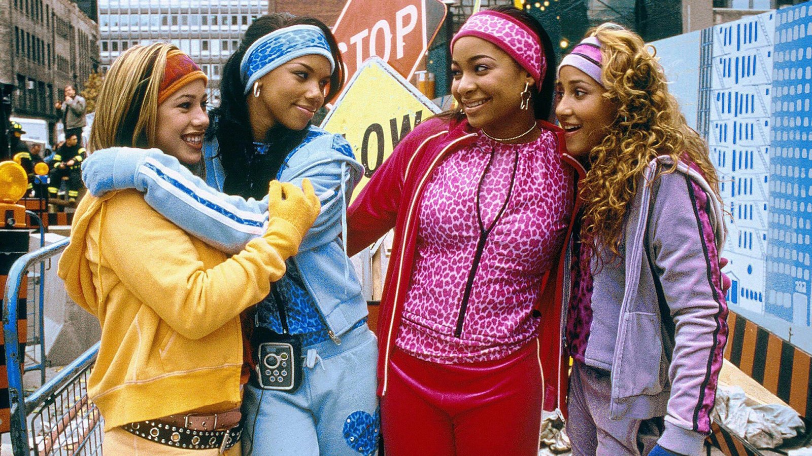 Quatre jeunes femmes vêtues de survêtements colorés se tiennent ensemble, souriantes, dans une rue de la ville, avec des panneaux de construction et des bâtiments en arrière-plan - canalisant l'énergie vibrante des Cheetah Girls d'un film classique de Disney Channel 4e.