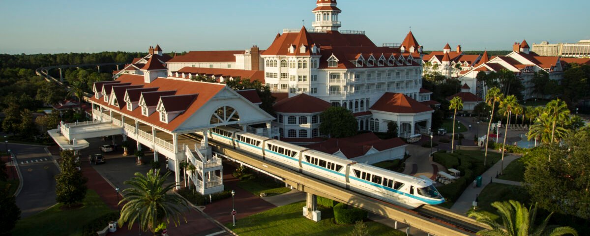 Un monorail blanc passe devant un grand hôtel à Walt Disney World, de style victorien avec des toits rouges et des murs blancs, entouré de palmiers et d'un parc paysager sous un ciel bleu clair.