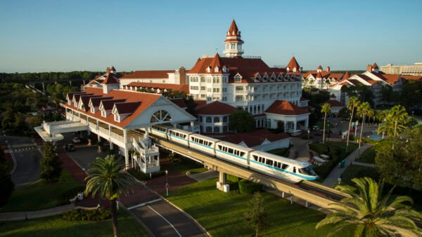 Un monorail blanc passe devant un grand hôtel à Walt Disney World, de style victorien avec des toits rouges et des murs blancs, entouré de palmiers et d'un parc paysager sous un ciel bleu clair.
