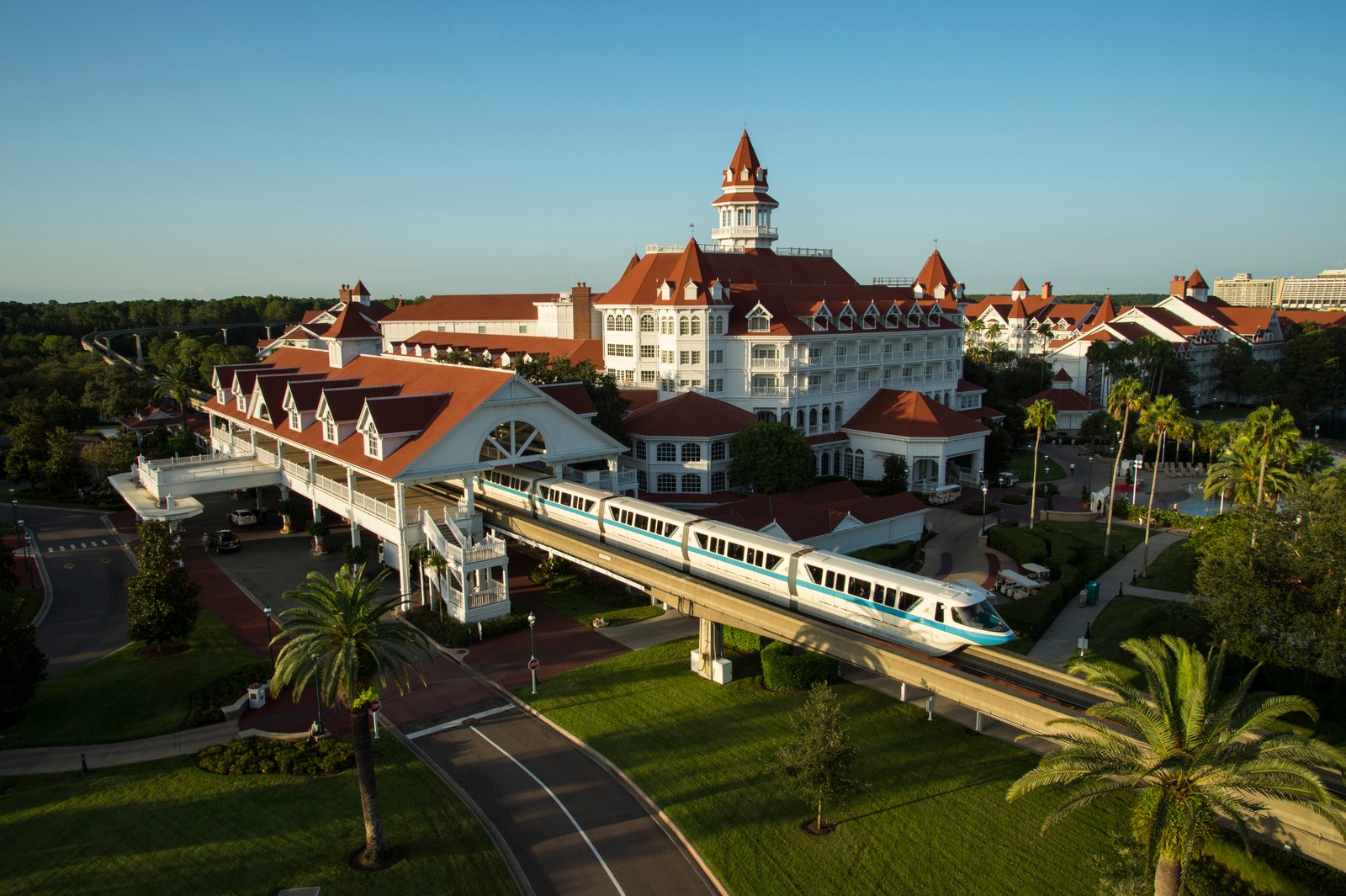 Un monorail blanc passe devant un grand hôtel à Walt Disney World, de style victorien avec des toits rouges et des murs blancs, entouré de palmiers et d'un parc paysager sous un ciel bleu clair.