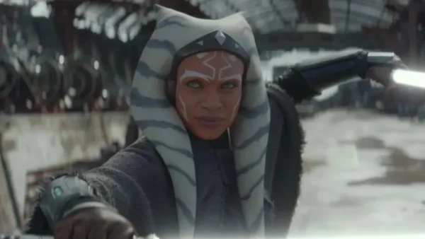 Une femme vêtue d'une cape grise avec des marques faciales blanches et une queue de pie rayée tient deux sabres laser lumineux dans une position défensive à l'intérieur d'une structure industrielle, laissant entrevoir des scènes d'Ahsoka Saison 2 de la série Star Wars, dont la sortie est prévue pour 2027.