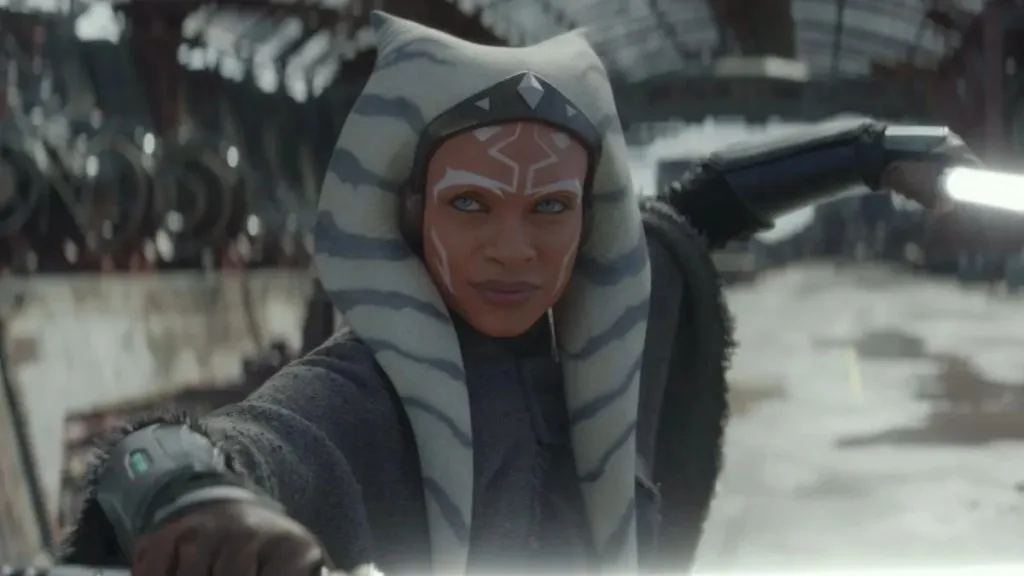 Une femme vêtue d'une cape grise avec des marques faciales blanches et une queue de pie rayée tient deux sabres laser lumineux dans une position défensive à l'intérieur d'une structure industrielle, laissant entrevoir des scènes d'Ahsoka Saison 2 de la série Star Wars, dont la sortie est prévue pour 2027.