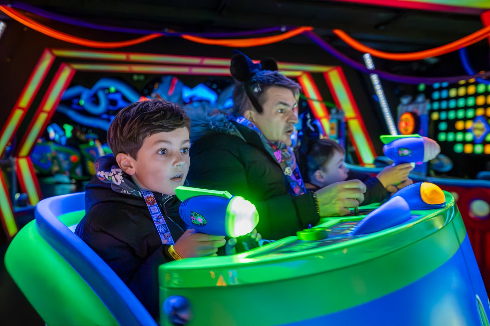 Un garçon et deux adultes se promènent dans une attraction de Disneyland Paris éclairée au néon, en visant des cibles avec des pistolets laser aux expressions concentrées, ce qui en fait l'une des meilleures attractions pour enfants.