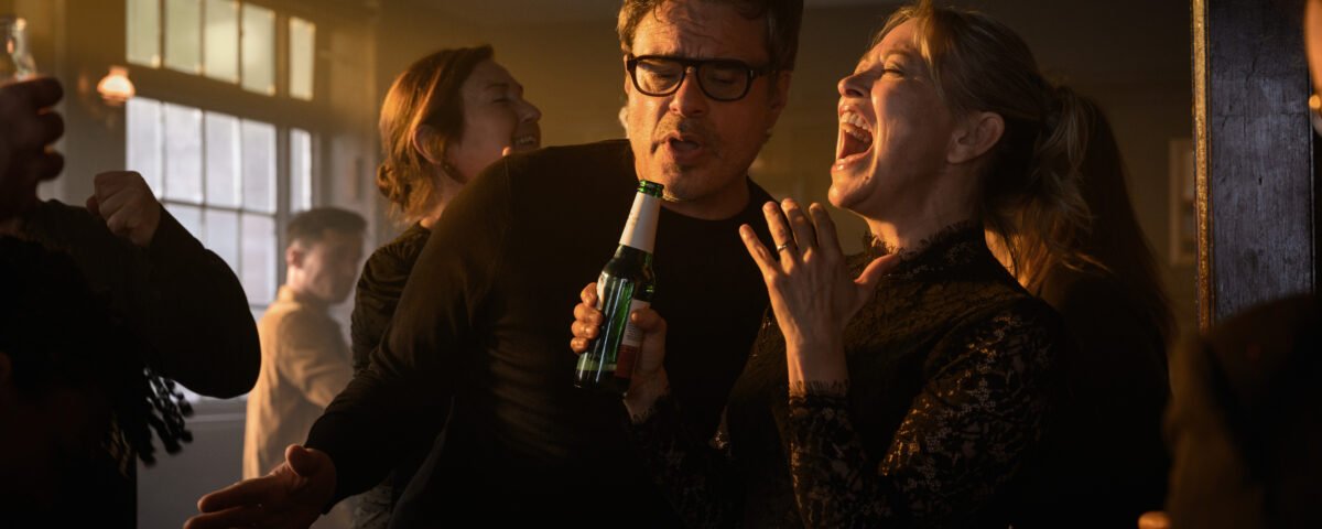 Deux personnes chantent ensemble dans un bar animé et faiblement éclairé, l'une tenant une bouteille de bière en guise de micro, canalisant l'esprit amusant d'Alice et Steve de la Comédie Hulu, tandis que d'autres personnes socialisent en arrière-plan.