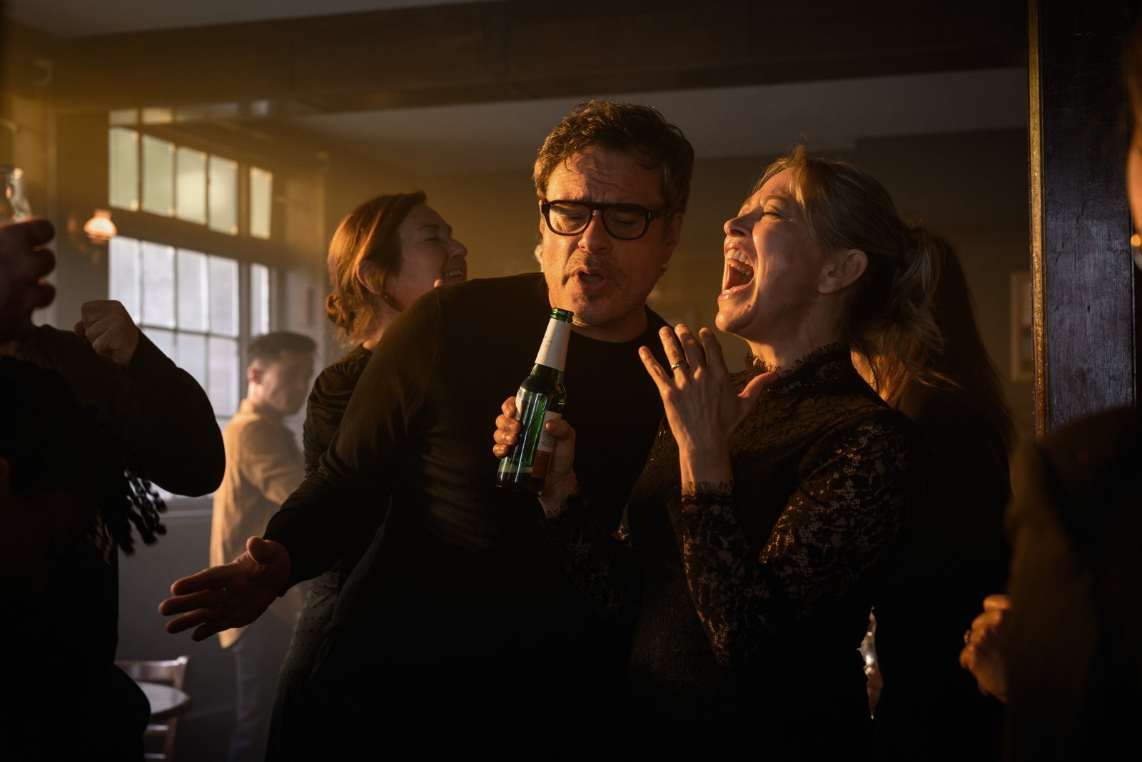 Deux personnes chantent ensemble dans un bar animé et faiblement éclairé, l'une tenant une bouteille de bière en guise de micro, canalisant l'esprit amusant d'Alice et Steve de la Comédie Hulu, tandis que d'autres personnes socialisent en arrière-plan.