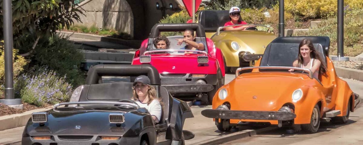 Quatre petites voitures Autopia, chacune avec des passagers, circulent en plein air dans une zone paysagée près de l'entrée d'un tunnel à Disneyland, mettant en valeur le charme des voitures à essence classiques.