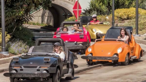 Quatre petites voitures Autopia, chacune avec des passagers, circulent en plein air dans une zone paysagée près de l'entrée d'un tunnel à Disneyland, mettant en valeur le charme des voitures à essence classiques.