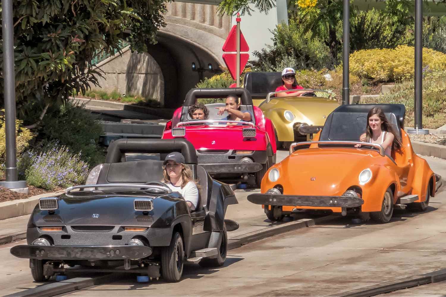 Quatre petites voitures Autopia, chacune avec des passagers, circulent en plein air dans une zone paysagée près de l'entrée d'un tunnel à Disneyland, mettant en valeur le charme des voitures à essence classiques.