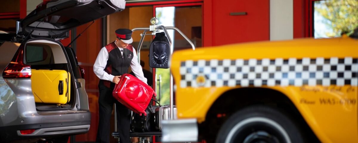 Un membre du personnel de l'hôtel charge les bagages sur un chariot à l'extérieur du Disney's Hotel New York - The Art of Marvel, tandis qu'un taxi jaune - idéal pour vérifier le Tarif Taxi Disneyland - et un SUV sont garés à proximité.