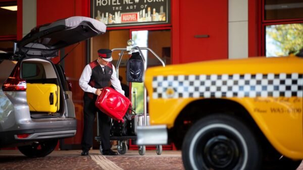 Un membre du personnel de l'hôtel charge les bagages sur un chariot à l'extérieur du Disney's Hotel New York - The Art of Marvel, tandis qu'un taxi jaune - idéal pour vérifier le Tarif Taxi Disneyland - et un SUV sont garés à proximité.