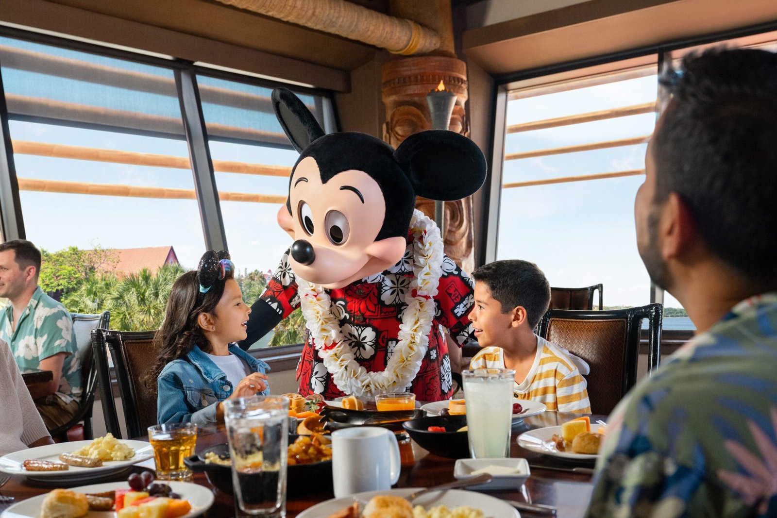 Une personne déguisée en Mickey Mouse et portant un lei floral interagit avec deux enfants à la table d'un restaurant, créant ainsi des expériences inoubliables qui rappellent la magie de Walt Disney World.