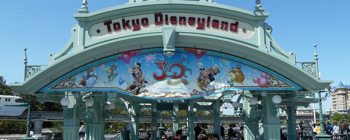 Porte d'entrée de Tokyo Disneyland avec des gens qui entrent, décorée de personnages et de drapeaux Disney, et un panneau célébrant le 30e anniversaire de l'ouverture du parc, une étape importante dans son histoire.