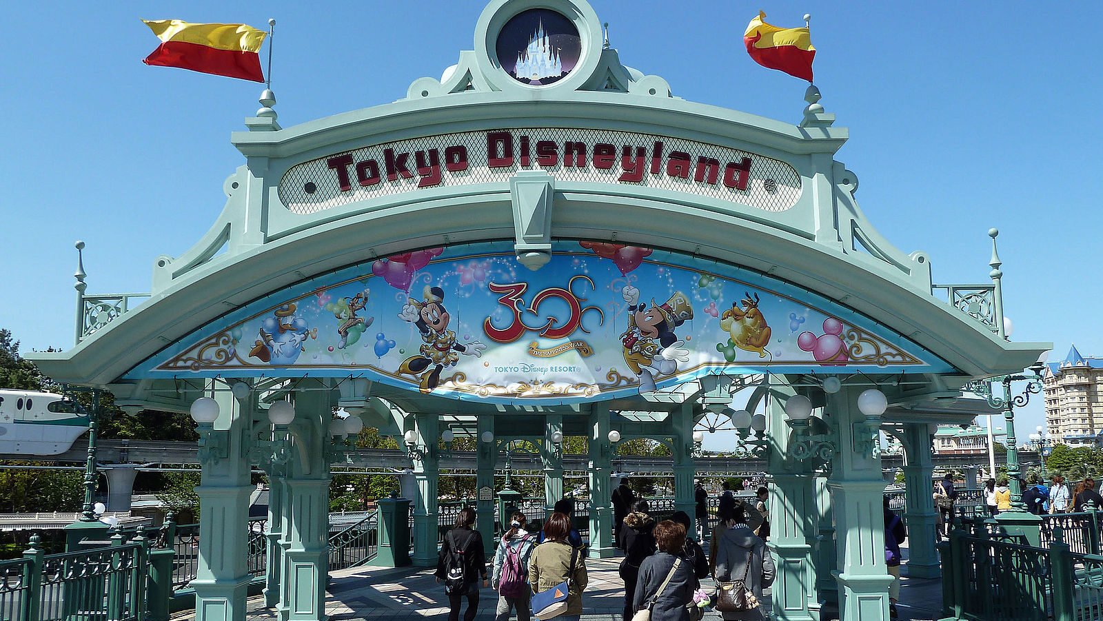 Porte d'entrée de Tokyo Disneyland avec des gens qui entrent, décorée de personnages et de drapeaux Disney, et un panneau célébrant le 30e anniversaire de l'ouverture du parc, une étape importante dans son histoire.
