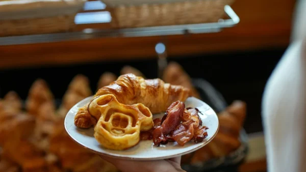 Au Disney's Davy Crockett Ranch, une personne tient une assiette avec des pâtisseries et du bacon devant des paniers remplis de pain et de croissants au Buffet Matinal.