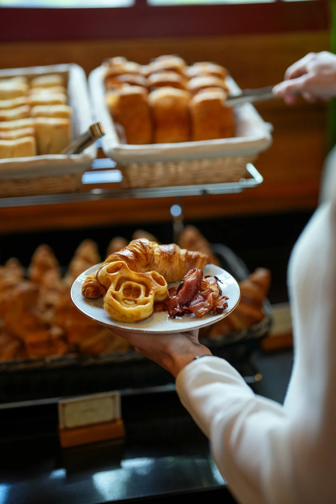 Au Disney's Davy Crockett Ranch, une personne tient une assiette avec des pâtisseries et du bacon devant des paniers remplis de pain et de croissants au Buffet Matinal.