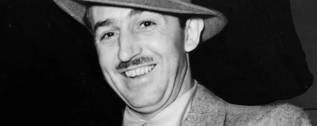Un homme en costume, cravate et chapeau sourit à la caméra sur un fond sombre, évoquant le charme classique de l'histoire de Disney.