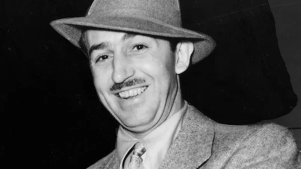 Un homme en costume, cravate et chapeau sourit à la caméra sur un fond sombre, évoquant le charme classique de l'histoire de Disney.