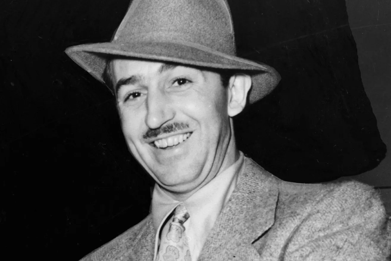Un homme en costume, cravate et chapeau sourit à la caméra sur un fond sombre, évoquant le charme classique de l'histoire de Disney.