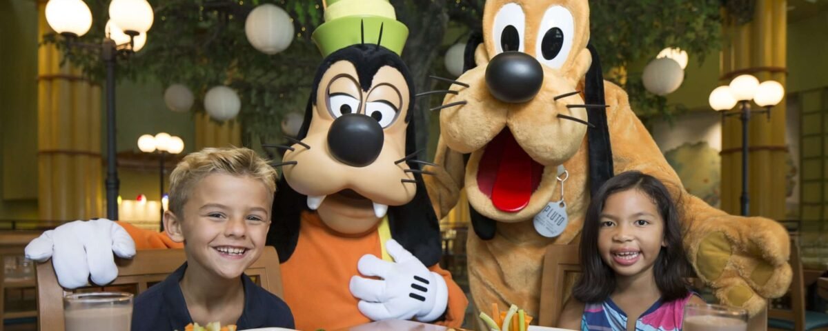 Deux enfants sont assis à une table avec de la nourriture et des boissons, rejoints par les personnages de Walt Disney World Goofy et Pluto en costume, créant ainsi des expériences inoubliables dans le cadre d'un restaurant illuminé.