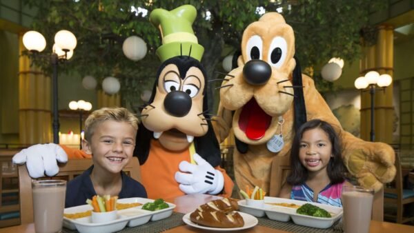 Deux enfants sont assis à une table avec de la nourriture et des boissons, rejoints par les personnages de Walt Disney World Goofy et Pluto en costume, créant ainsi des expériences inoubliables dans le cadre d'un restaurant illuminé.