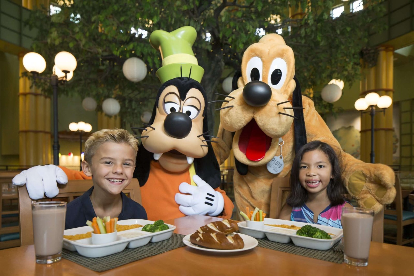 Deux enfants sont assis à une table avec de la nourriture et des boissons, rejoints par les personnages de Walt Disney World Goofy et Pluto en costume, créant ainsi des expériences inoubliables dans le cadre d'un restaurant illuminé.