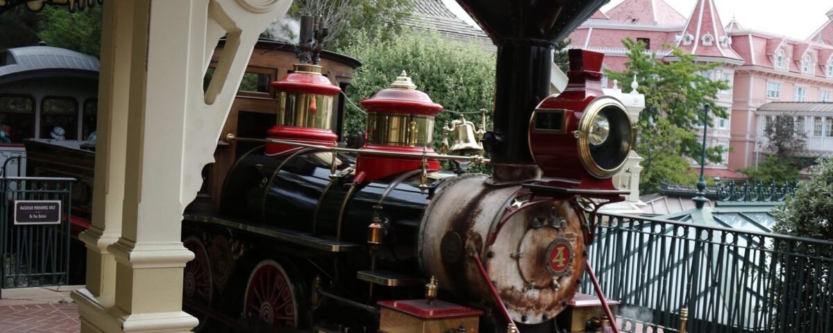 Une locomotive à vapeur d'époque portant le numéro 4 à l'avant se trouve sur le quai d'une gare, dégageant de la vapeur, avec des bâtiments de style victorien en arrière-plan - une scène charmante qui rappelle les transports et l'accessibilité de Disneyland Paris.