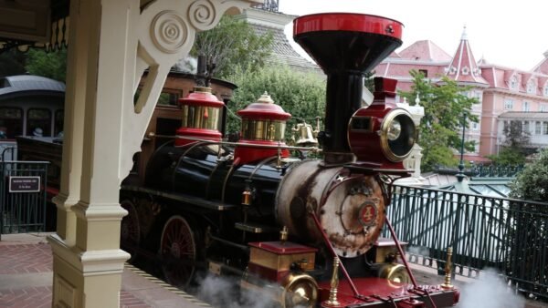 Une locomotive à vapeur d'époque portant le numéro 4 à l'avant se trouve sur le quai d'une gare, dégageant de la vapeur, avec des bâtiments de style victorien en arrière-plan - une scène charmante qui rappelle les transports et l'accessibilité de Disneyland Paris.