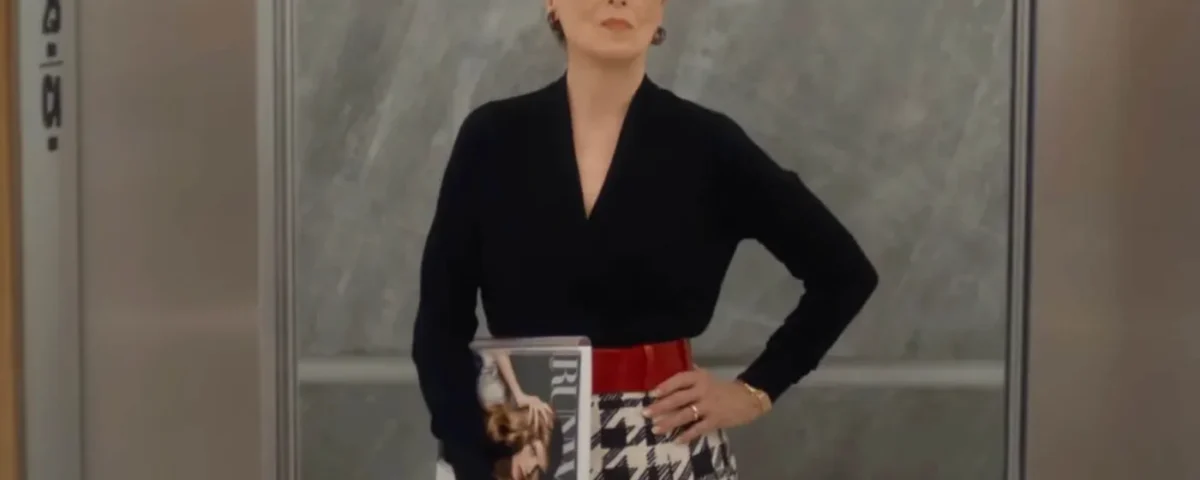 Une femme aux cheveux blancs et aux lunettes de soleil se tient dans un ascenseur, un magazine à la main, vêtue d'un haut noir, d'une jupe pied-de-poule et d'une ceinture rouge - un look qui rappelle le succès mondial du Diable S'habille En Prada au box-office.