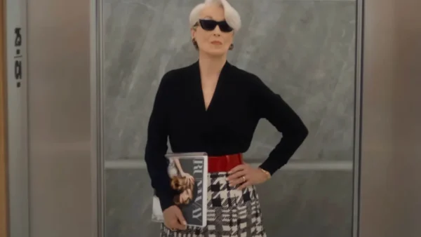 Une femme aux cheveux blancs et aux lunettes de soleil se tient dans un ascenseur, un magazine à la main, vêtue d'un haut noir, d'une jupe pied-de-poule et d'une ceinture rouge - un look qui rappelle le succès mondial du Diable S'habille En Prada au box-office.