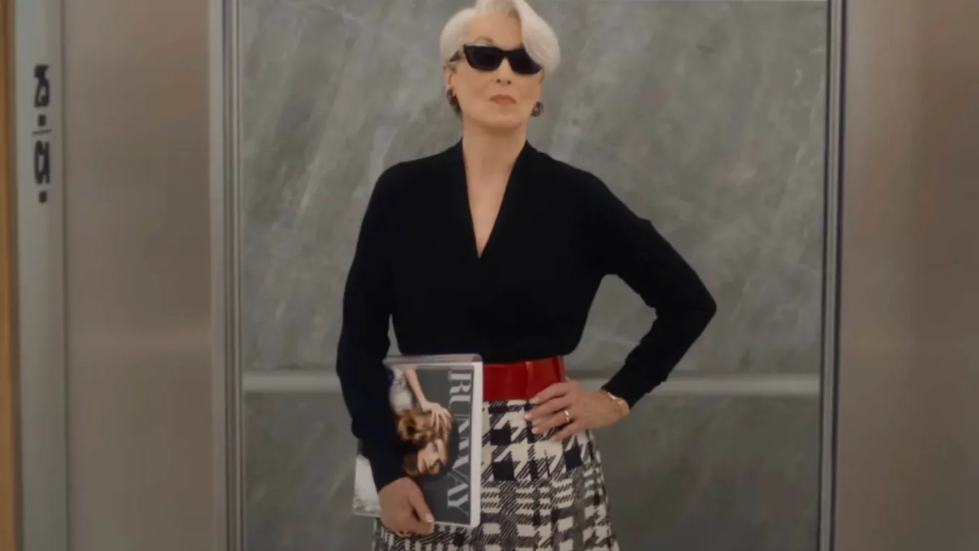 Une femme aux cheveux blancs et aux lunettes de soleil se tient dans un ascenseur, un magazine à la main, vêtue d'un haut noir, d'une jupe pied-de-poule et d'une ceinture rouge - un look qui rappelle le succès mondial du Diable S'habille En Prada au box-office.