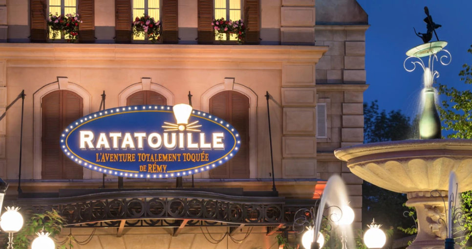 Une façade de bâtiment avec un panneau "Ratatouille" éclairé en français et une fontaine voisine surmontée d'une petite statue de rat, photographiée à la tombée de la nuit à l'attraction Ratatouille, qui a ouvert ses portes le 10 juillet 2014.